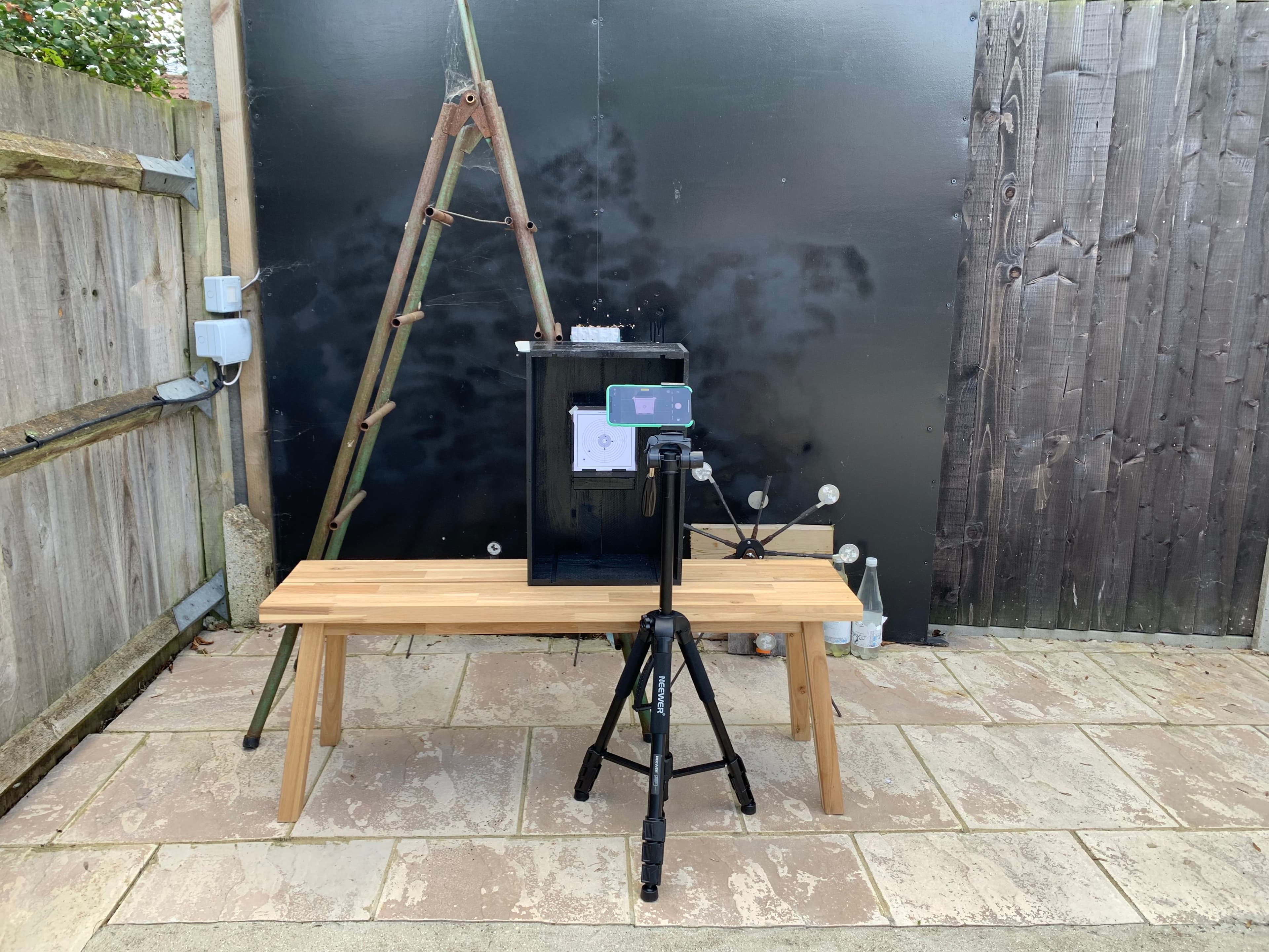 Backyard test rig — wooden bench, phone on tripod, target box against black backstop
